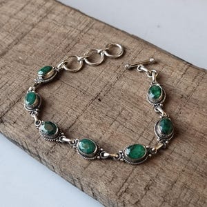 May include: A silver bracelet with a toggle clasp features a row of oval-shaped green gemstones. The bracelet is displayed on a wooden surface.