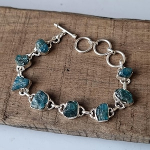 May include: A silver bracelet with a toggle clasp features a chain with eight blue-green gemstone links. The gemstones are rough and irregular in shape.