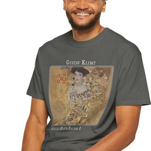 May include: A charcoal gray t-shirt featuring a portrait of Adele Bloch-Bauer I by Gustav Klimt. The artwork is in a gold and brown color palette. The text "GUSTAV KLIMT" and "ADELE BLOCH-BAUER I" are printed above and below the portrait.