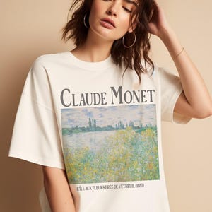 May include: Cream-colored t-shirt featuring the text "Claude Monet" above a landscape painting. The artwork depicts a field of flowers near a body of water. The shirt has short sleeves and a relaxed fit.