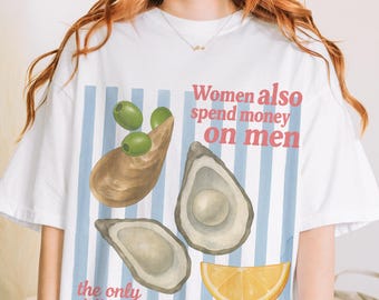 以下が含まれることがあります： カキ、オリーブ、レモンスライスをあしらったグラフィックデザインの白いTシャツ。「女性も男性にお金を使いますが、唯一の違いは女性が騒がないことです」と書かれています。