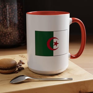 Könnte beinhalten: Ein weißer Keramikbecher mit der aufgedruckten Nationalflagge von Algerien auf der Vorderseite. Der Becher hat ein leuchtend rotes Inneres und einen passenden roten Henkel. Das Flaggendesign besteht aus einem grün-weißen vertikalen Bicolor.