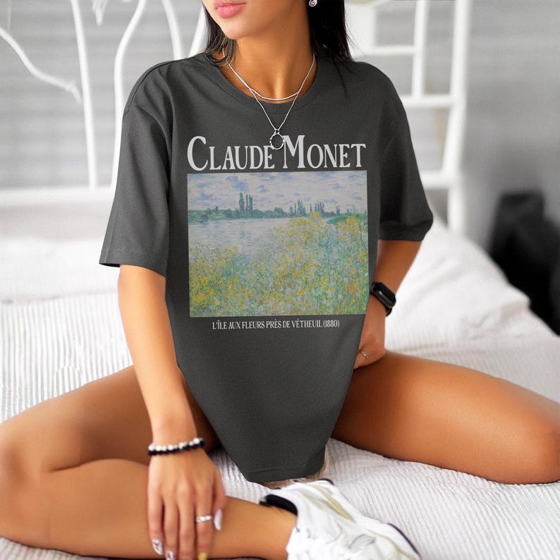 May include: Dark gray t-shirt featuring a Claude Monet painting, with the artist's name in white above the artwork. The artwork depicts a landscape with a river and yellow flowers. The text "L'ILE AUX FLEURS PRES DE VETHEUIL (1880)" is below the painting.