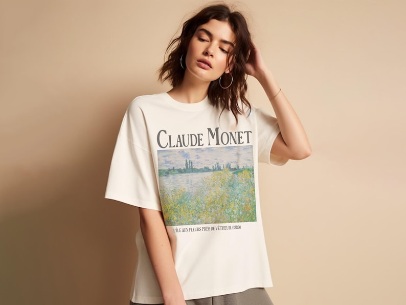 May include: Cream-colored t-shirt featuring the text "Claude Monet" above a landscape painting. The artwork depicts a field of flowers near a body of water. The shirt has short sleeves and a relaxed fit.