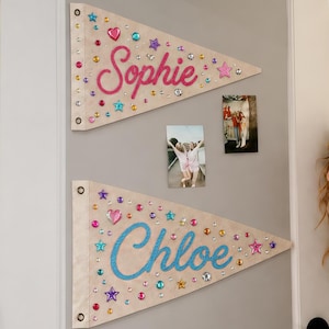 May include: Two triangular beige fabric pennants featuring the names Sophie and Chloe in cursive script. The pennants are decorated with colourful plastic gems in shapes of hearts, stars, and circles. Each pennant has two metal grommets on the left side.