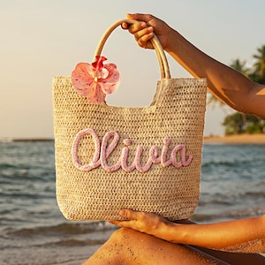 May include: A woven straw tote bag with the name "Olivia" in pink script. The bag has a bamboo handle and a pink flower accent. The background shows a beach scene with palm trees and ocean.