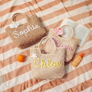 May include: Three woven straw tote bags with handles, each personalised with a name in a different colour: "Sophie" in white, "Olivia" in pink, and "Chloe" in yellow. The bags are on a striped beach towel.