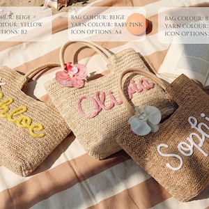 May include: Three woven straw tote bags with embroidered names: "Chloe" in yellow, "Olivia" in baby pink, and "Sophie" in white. Each bag has a wooden handle and is decorated with a flower. The bags are on a striped surface.