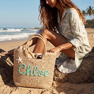May include: A woven straw tote bag with the name "Chloe" in turquoise script. A white starfish is attached to the bag. The bag has two handles and is sitting on a sandy beach.