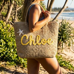 May include: A woven straw tote bag with the name "Chloe" in yellow script. A white starfish is attached to the bag. The bag is carried on a beach with a blue ocean in the background.