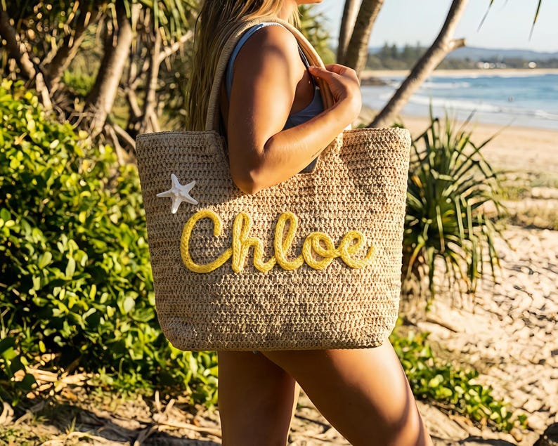 May include: A woven straw tote bag with the name "Chloe" in yellow script. A white starfish is attached to the bag. The bag is carried on a beach with a blue ocean in the background.