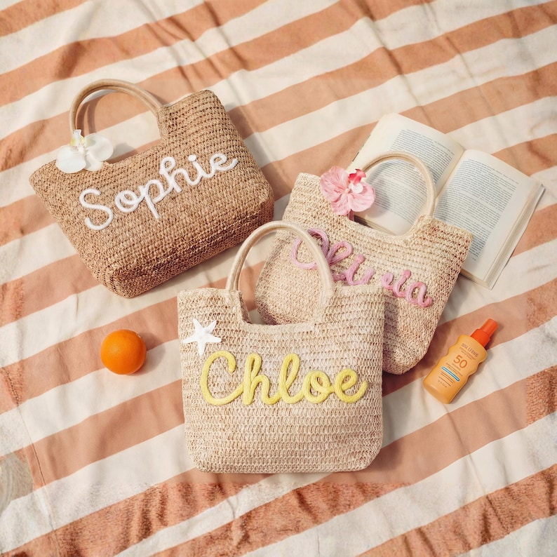 May include: Three woven straw tote bags with handles, each personalized with a name in a different color: "Sophie" in white, "Olivia" in pink, and "Chloe" in yellow. The bags are on a striped beach towel.
