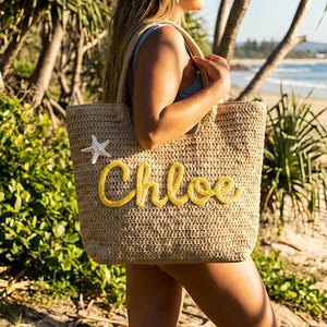 May include: A beige woven tote bag with the name "Chloe" in yellow script. A white starfish is attached to the bag. The bag has a rectangular shape and is being held by a person.