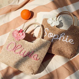 May include: Two woven straw tote bags with handles, one with "Olivia" embroidered in pink and a pink flower, the other with "Sophie" in white and a white flower. An orange and a book are on a striped beach towel.