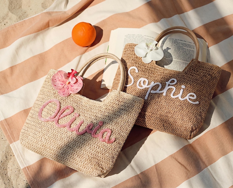 May include: Two woven straw tote bags with handles, one with "Olivia" embroidered in pink and a pink flower, the other with "Sophie" in white and a white flower. An orange and a book are on a striped beach towel.