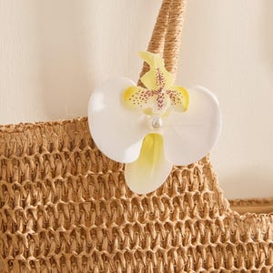 May include: A woven straw tote bag with a decorative white and yellow orchid flower attached to the handle. The orchid has a pearl-like center and is accented with brown spots. The bag is a natural beige color.