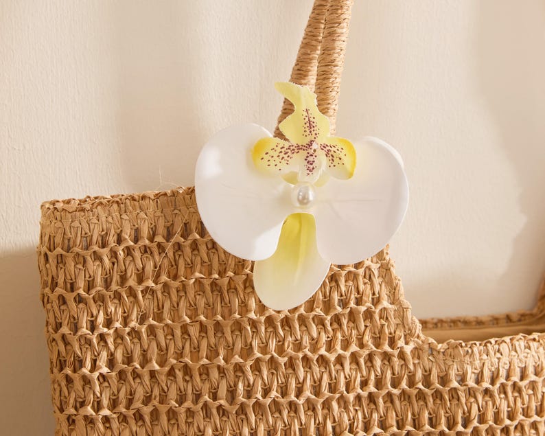 May include: A woven straw tote bag with a decorative white and yellow orchid flower attached to the handle. The orchid has a pearl-like center and is accented with brown spots. The bag is a natural beige color.