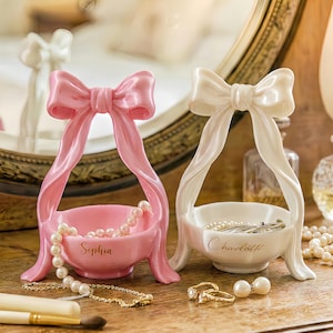 May include: Two decorative jewelry dishes, one pink and one white, each shaped like a bow with a small bowl. The pink dish has the name "Sophia" and the white dish has the name "Charlotte" inscribed on them. Both are filled with jewelry.