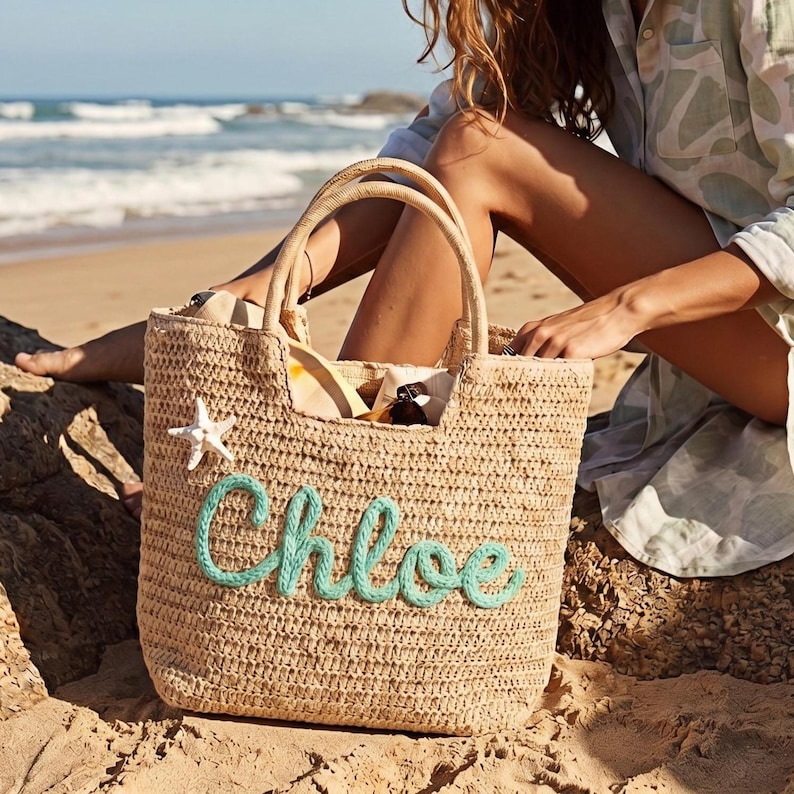 May include: A woven straw tote bag with the name "Chloe" in turquoise script. The bag has two handles and a white starfish accent. The bag is filled with items and is sitting on a rock near the ocean.