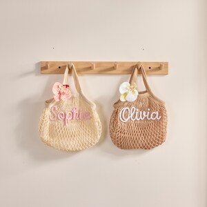 May include: Two beige net tote bags, one with the name "Sophie" in pink script and a pink flower, the other with "Olivia" in white script and a white flower. Both bags hang from a wooden rack.