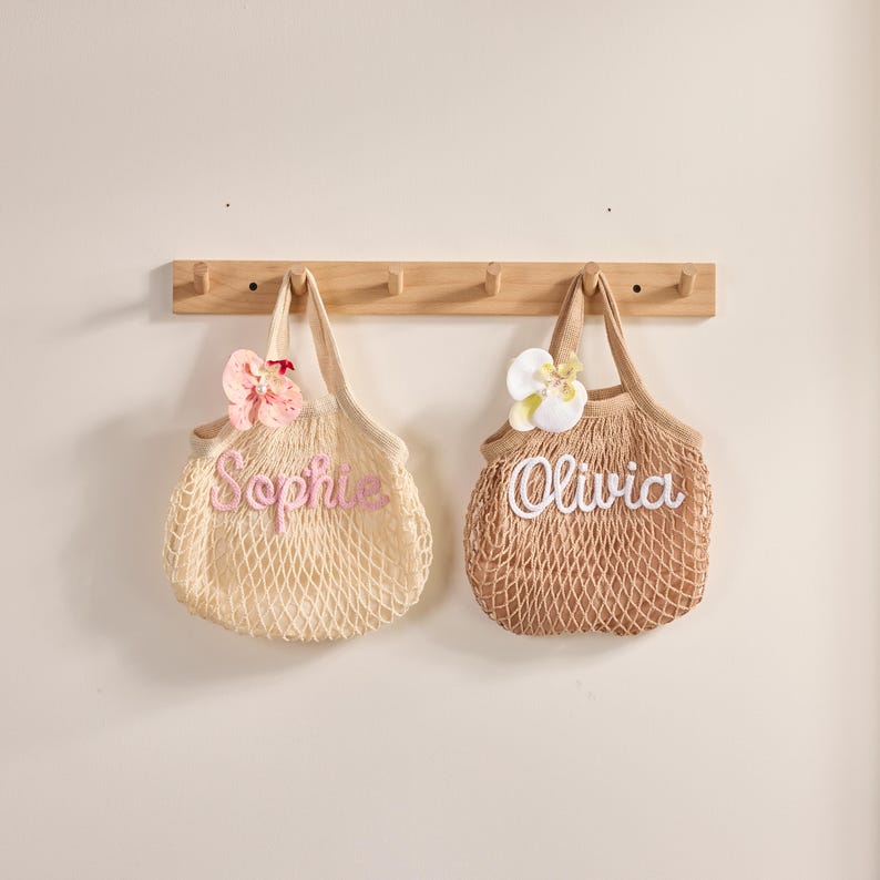 May include: Two beige net tote bags, one with the name "Sophie" in pink script and a pink flower, the other with "Olivia" in white script and a white flower. Both bags hang from a wooden rack.