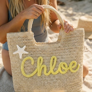 May include: A woven straw tote bag with a cream-colored starfish and the name "Chloe" in yellow script. The bag has a rectangular shape with a handle and is perfect for carrying beach essentials. The bag is made of natural materials.