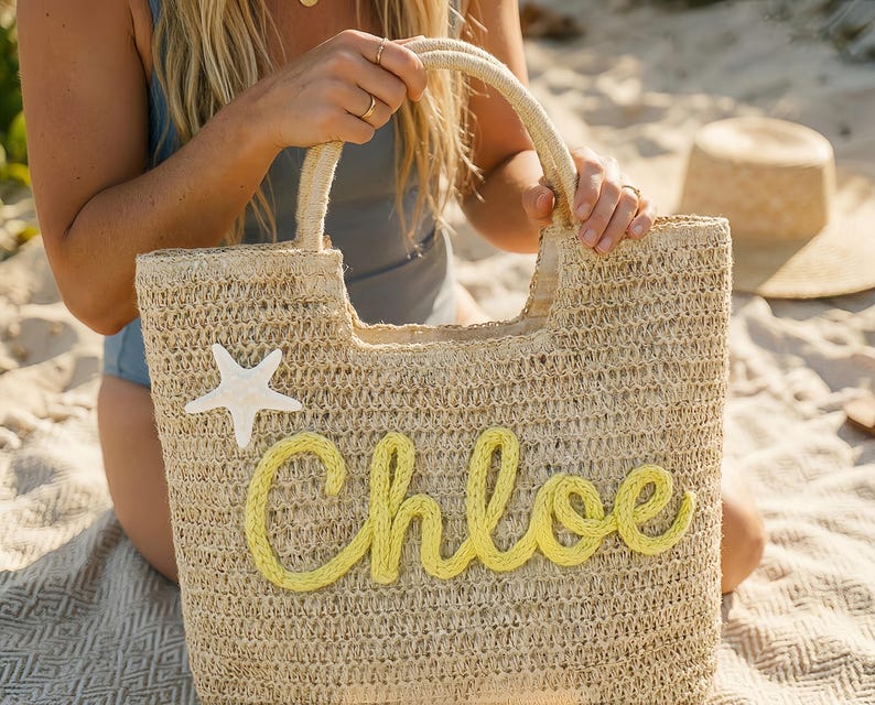 May include: A woven straw tote bag with a cream-colored starfish and the name "Chloe" in yellow script. The bag has a rectangular shape with a handle and is perfect for carrying beach essentials. The bag is made of natural materials.