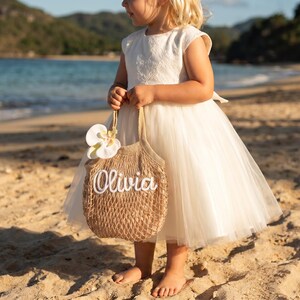 May include: A young girl in a white dress holds a woven tote bag with the name "Olivia" in white script. The bag is adorned with a white flower. The scene is set on a sandy beach with the ocean in the background.