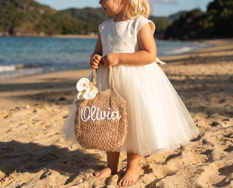 May include: A young girl in a white dress holds a woven tote bag with the name "Olivia" in white script. The bag is adorned with a white flower. The scene is set on a sandy beach with the ocean in the background.