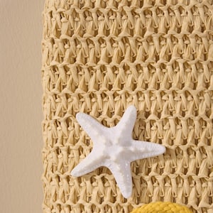 May include: Close-up of a woven straw bag with a white starfish accent. The bag has a textured, beige pattern. The starfish is white and has a textured surface. The bag is a beach or summer-themed accessory.