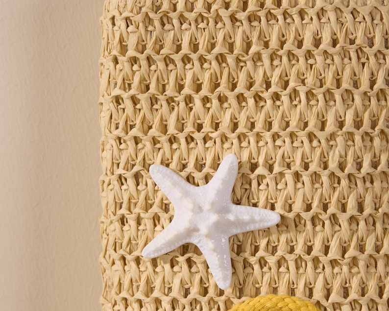 May include: Close-up of a woven straw bag with a white starfish accent. The bag has a textured, beige pattern. The starfish is white and has a textured surface. The bag is a beach or summer-themed accessory.