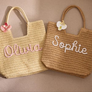 May include: Two woven straw tote bags. One is light yellow with the name "Olivia" in pink script. The other is tan with the name "Sophie" in white script. Both bags have handles and decorative floral accents.