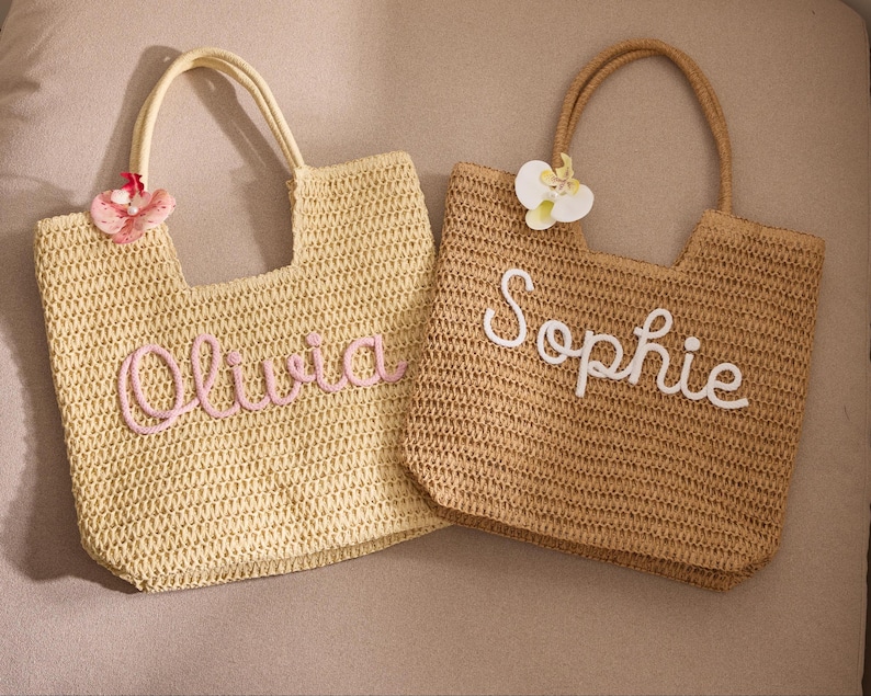 May include: Two woven straw tote bags. One is light yellow with the name "Olivia" in pink script. The other is tan with the name "Sophie" in white script. Both bags have handles and decorative floral accents.