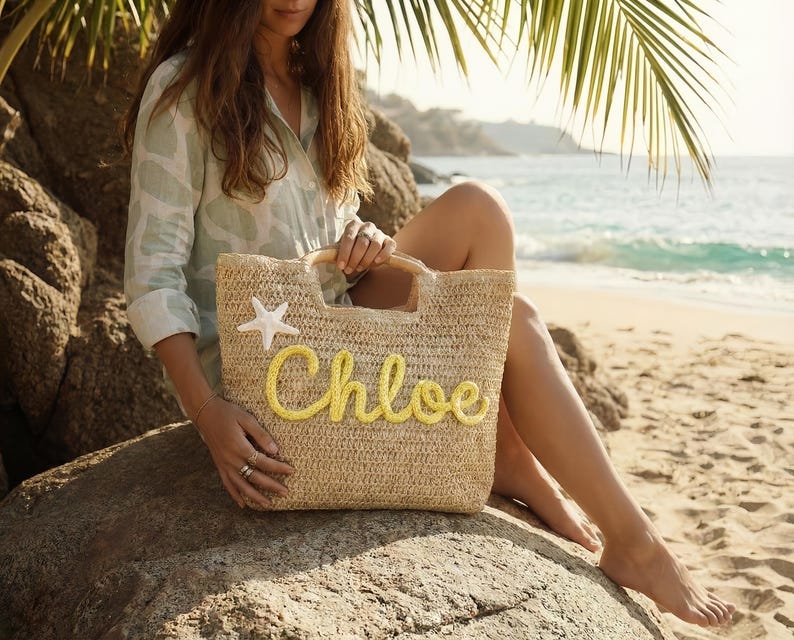 May include: A woven straw tote bag with wooden handles and the name "Chloe" in yellow script. A white starfish is attached to the bag. The bag is on a rock near a beach with palm fronds and ocean in the background.
