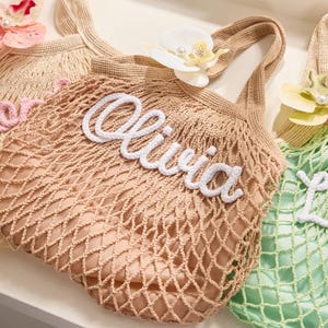 May include: Three crocheted net bags in beige, tan, and green, each with a name in white or pink script. The bag in the center says "Olivia". Each bag has a flower attached to the handle. The bags are designed for carrying items.