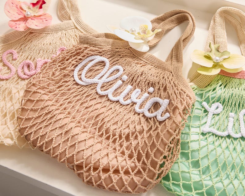 May include: Three crocheted net bags in beige, tan, and green, each with a name in white or pink script. The bag in the center says "Olivia". Each bag has a flower attached to the handle. The bags are designed for carrying items.