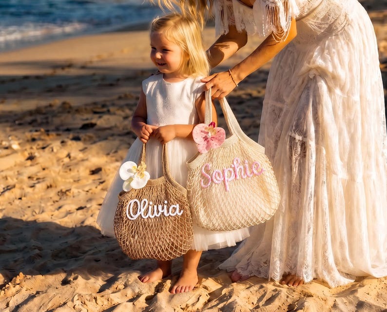 May include: Two mesh tote bags, one brown and one cream, held by a young girl on a beach. The brown bag has the name "Olivia" in white script, and the cream bag has "Sophie" in pink script. Both bags are decorated with floral accents.