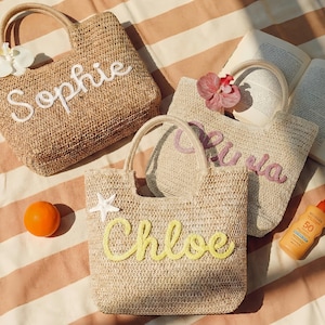 May include: Three woven straw tote bags with handles, each personalized with a name in a cursive font: "Sophie", "Olivia", and "Chloe". The bags are decorated with a flower, a starfish, and a small orange. A bottle of sunscreen and an open book are also visible.