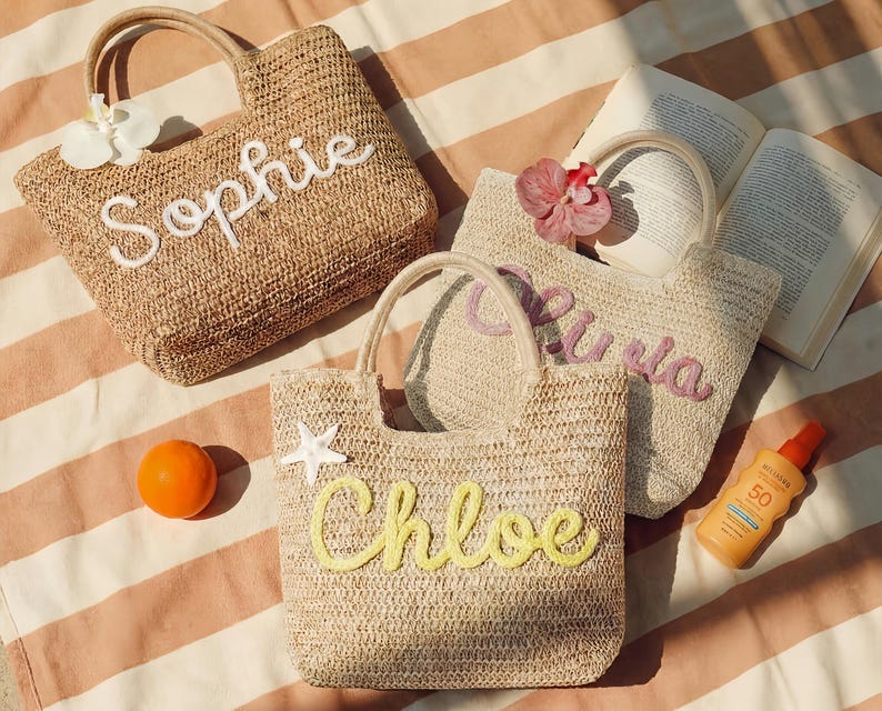 May include: Three woven straw tote bags with handles, each personalized with a name in a cursive font: "Sophie", "Olivia", and "Chloe". The bags are decorated with a flower, a starfish, and a small orange. A bottle of sunscreen and an open book are also visible.