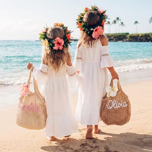 May include: Two young girls wearing white dresses and flower crowns walk on a sandy beach. Each girl carries a beige tote bag with a name embroidered on it. The ocean and palm trees are visible in the background.