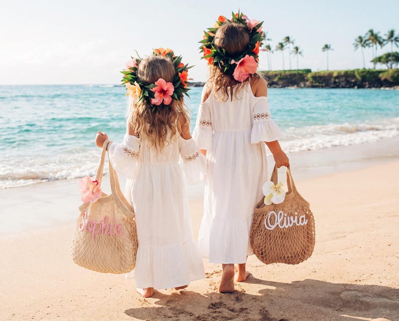 May include: Two young girls wearing white dresses and flower crowns walk on a sandy beach. Each girl carries a beige tote bag with a name embroidered on it. The ocean and palm trees are visible in the background.
