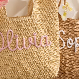 May include: Two woven straw tote bags. One is light tan with the name "Olivia" in pink script. The other is brown with the name "Soph" in white script. Both bags have floral embellishments and woven handles.