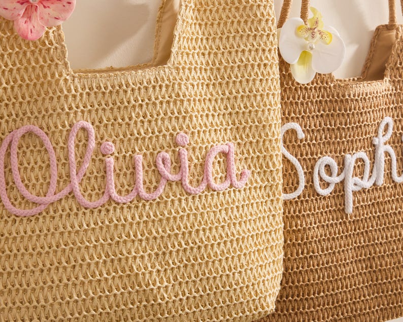 May include: Two woven straw tote bags. One is light tan with the name "Olivia" in pink script. The other is brown with the name "Soph" in white script. Both bags have floral embellishments and woven handles.
