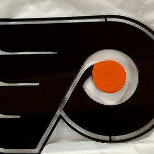 May include: Black and orange metal wall art featuring the Philadelphia Flyers logo. The logo is a stylized letter "P" with a wing on either side.