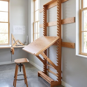 Op de afbeelding: Een lichtbruin houten wandbureau met een verstelbaar schuin werkvlak en een verticaal steunframe met inkepingen. Een bijpassende houten kruk met een donker metalen onderstel en een ronde voetensteun staat op de vloer.