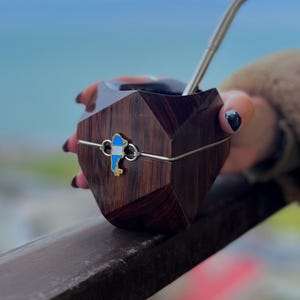 May include: A dark wooden yerba mate cup with a geometric design, featuring a silver rim and a decorative emblem with blue and white accents. A silver straw is inserted into the cup. The cup is held by a person.