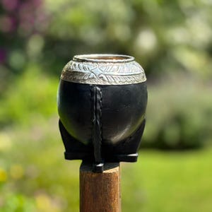 May include: A black gourd-shaped yerba mate cup with a silver-colored metal rim featuring a leaf design. The cup is mounted on a wooden post against a blurred green background. The cup has a stitched seam down the back.