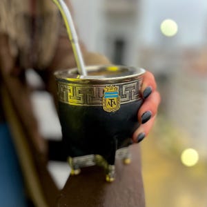 Puede incluir: Un mate negro con borde y base plateados, con un emblema con las letras "AFA" y un diseño azul y amarillo. Una pajita plateada se extiende desde la parte superior. El mate es sostenido por una persona.