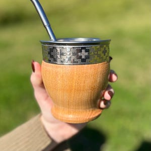 Puede incluir: Una calabaza de yerba mate de madera con un borde y bombilla de acero inoxidable. El borde presenta un patrón decorativo. La calabaza se sostiene frente a un fondo verde borroso.