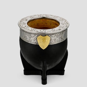 Puede incluir: Una calabaza de yerba mate negra con borde y banda decorativa plateados. Un escudo dorado con las letras "CABJ" está fijado en la parte delantera. La calabaza descansa sobre una base negra con tres pequeños pies. Aproximadamente 10 cm de altura.